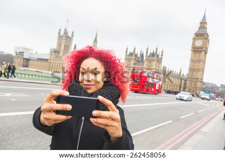 Smiling red hair girl taking selfie in London with Big Ben on background. She is holding the phone and looking at camera. Also there is a red double decker bus on the street behind her.