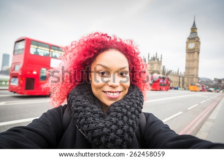Smiling red hair girl taking selfie in London with Big Ben on background. She is holding the phone and looking at camera. Also there is a red double decker bus on the street behind her.