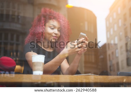 Beautiful girl with curly red hair typigin on smart phone in a cafe, seen through the window with buildings reflections