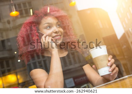 Beautiful girl with curly red hair talking on smart phone in a cafe, seen through the window with buildings reflections