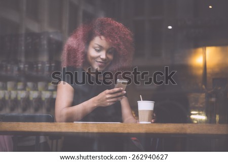 Beautiful girl with curly red hair typigin on smart phone in a cafe, seen through the window with buildings reflections
