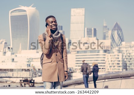 Young black man talking on mobile phone in London with city skyscrapers on background in a sunny day. He is wearing a coat and has a vintage backpack. Vintage filter applied.
