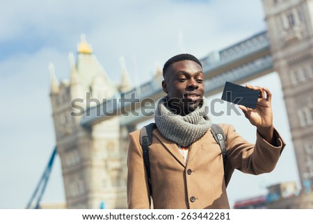 Smiling black man taking selfie in London with Tower Bridge on background. He is holding the phone and looking at camera. Photo taken on a sunny winter day.Vintage filter applied.