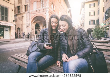 Two female twins looking at a smart phone. They wear black jackets and jeans. Urban scene with houses and street on background.