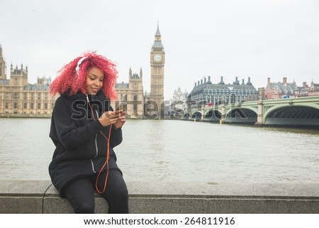 Beautiful redhair woman listening music in London with Big Ben and Westminster palace on background. She wear fashion white headphones.