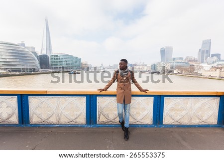 Young black man standing on Tower Bridge with London skyline  on background. He wears a brown coat and light blue jeans.