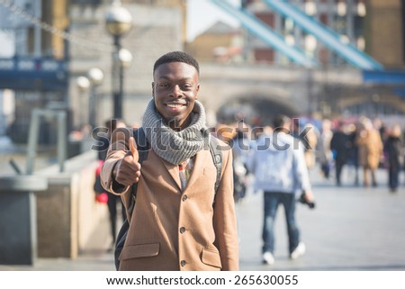 Successful young black man in London on Thames sidewalk showing thumbs up, with Tower Bridge and blurred people on background.