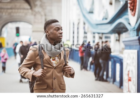 Young black man in London walking  on Tower Bridge. He wears a brown coat and light blue jeans. Traffic and other persons on background.vintage filter applied.