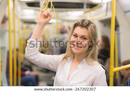 Beautiful blonde young woman holding with right hand inside tube train in London. She wears a white shirt and she is looking at camera smiling.