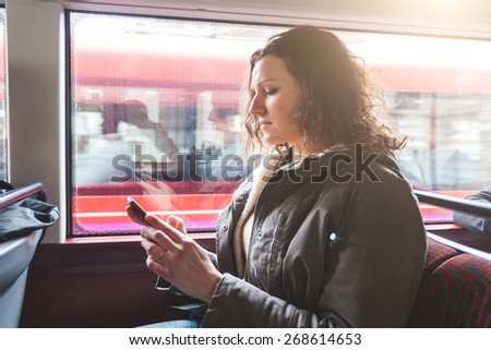 Beautiful woman typing on smart phone while commuting in London with double-decker bus. She is sitting next to the window with buildings and traffic on background.