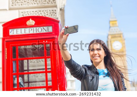 Beautiful young woman in London taking a selfie with Big Ben and red phone booth on background. She is holding the phone with right hand and looking at it. Focus on the face.
