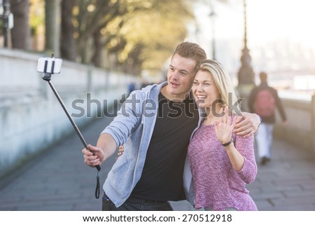 Young couple taking a selfie with smartphone on the stick. The man is holding the stick and embracing the girl, she is waving while looking at phone. Backlight flare added.