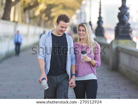 Young couple walking in London, talking and smiling, and holding a cup of tea. They are in their twenties, wearing light clothing and holding hands.
