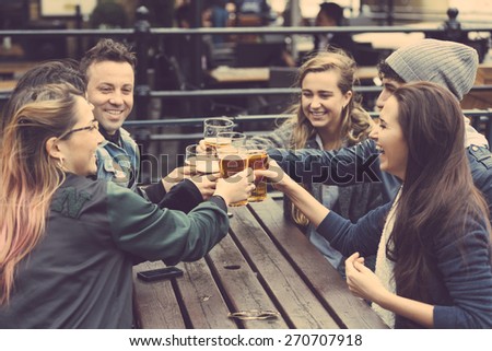 Group of friends enjoying a beer at pub in London, toasting and laughing. They are four girls and two boys in their twenties.