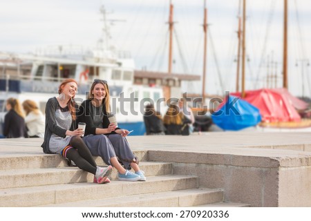 Two beautiful nordic girls at Oslo harbour enjoying life, talking and looking at smart phone, with ships on background. They also hold a cup of coffee in the hands. Lifestyle and friendship concepts.
