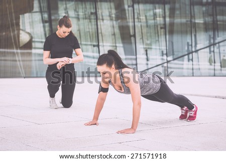 Woman doing push-ups exercises with her personal trainer in a modern urban context. She is wearing gray and black sportswear and a phone holder, the trainer look at watch and incite her