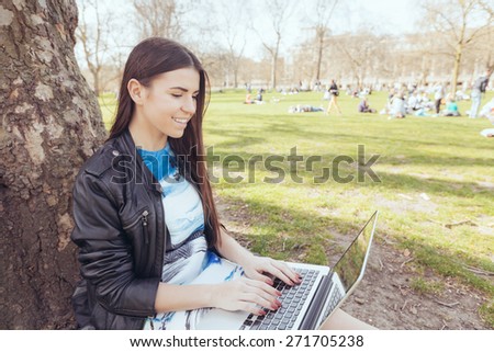 Young woman using computer at park in London. She is lying next to a tree, resting in the shadow on a London sunny day in spring.