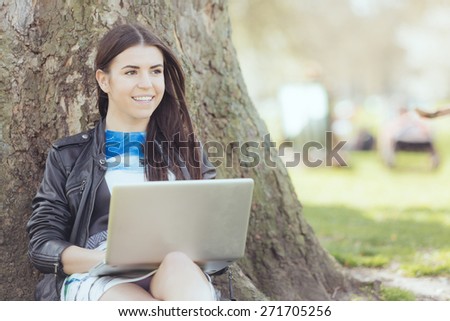 Young woman using computer at park in London. She is lying next to a tree, resting in the shadow on a London sunny day in spring.
