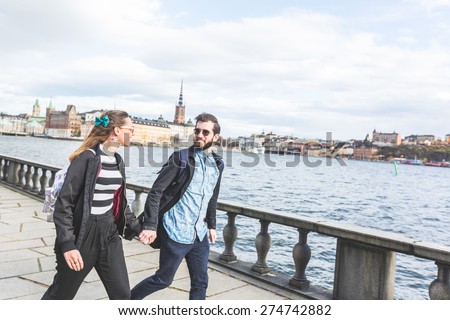 Young hipster couple running hand in hand in Stockholm with sea and old town on background. They are both wearing sunglasses and laughing while running together.