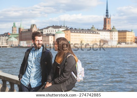 Young hipster couple visiting Stockholm. They are walking with sea and old town on background. Both are wearing sunglasses and a black jacket.