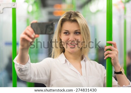Beautiful blonde young woman taking a selfie with smart phone in London tube train. She is holding to a pole, wearing a white shirt and looking at the phone.