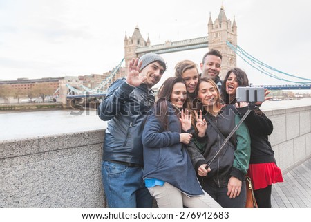 Group of friends taking a selfie using a selfie stick in London with Tower Bridge on background. They are four girls and two boys in their twenties, embracing and having fun together.