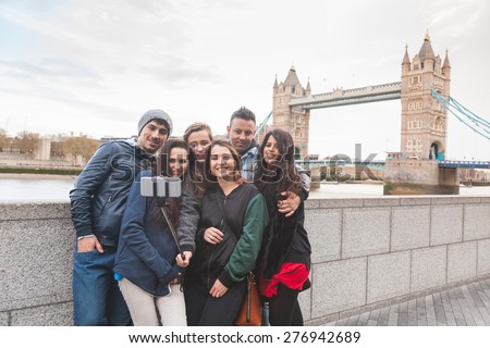 Group of friends taking a selfie using a selfie stick in London with Tower Bridge on background. They are four girls and two boys in their twenties, embracing and having fun together.