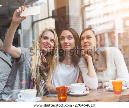 Happy women taking a selfie in a cafe in Copenhagen. They are in their twenties, enjoying a cup of coffee or tea and wearing smart casual clothes.