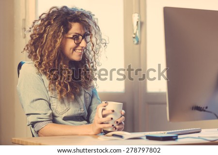 Young woman working at home or in a small office, vintage hipster clothing, curly hair. Cup of tea or coffee on the desk with some technological devices.