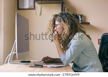 Young woman working at home or in a small office, vintage hipster clothing, curly hair. On the wooden desk there are a computer, a digital tablet, a smart phone and a notepad.