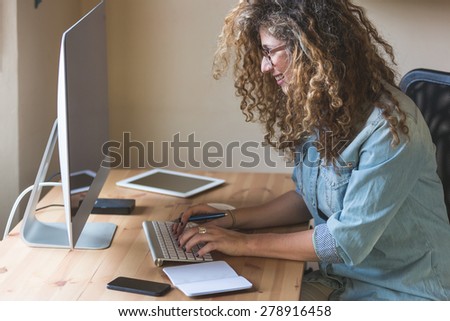 Young woman working at home or in a small office, vintage hipster clothing, curly hair. On the wooden desk there are a computer, a digital tablet, a smart phone and a notepad.