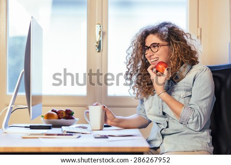 Young woman working at home or in a small office, vintage hipster clothing, curly hair. She is eating some fresh fruits, there is a cup of tea or coffee on the desk with some technological devices.