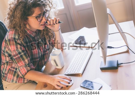 Young woman working at home or in a small office, vintage hipster clothing, curly hair. She seems to be thoughtful, There is a cup of tea or coffee on the desk with some technological devices