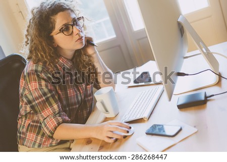 Young woman working at home or in a small office, vintage hipster clothing, curly hair. She seems to be thoughtful, There is a cup of tea or coffee on the desk with some technological devices