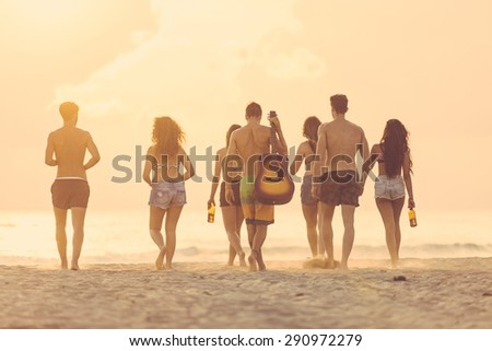 Group of friends walking on the beach at sunset. There are four girls and three boys, the sand is blowing, backlight technique, rear view.