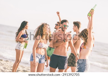 Multiracial group of friends having a party on the beach, dancing and drinking beer. They are teenagers, four girls and three boys, standing just next to the seaside.