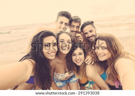 Multiracial group of friends taking selfie on the beach, camera point of view. They are teenagers, four girls and three boys, standing just next to the seaside.