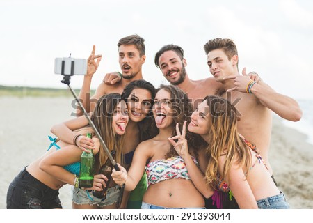 Multiracial group of friends taking selfie with a selfie stick on the beach. They are teenagers, four girls and three boys, standing just next to the seaside.