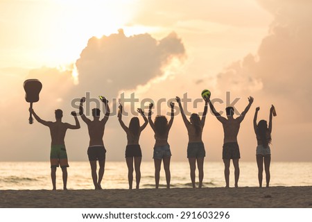Silhouette of a group of friends with raised hands on the beach at sunset. There are four girls and three boys, one is holding a guitar and another one a ball. Backlight technique.