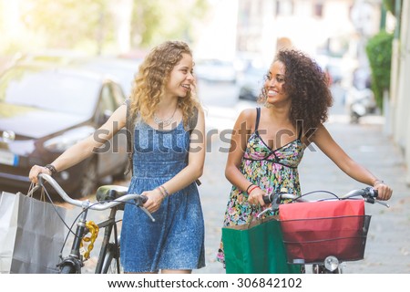 Multiracial couple riding bikes on the street. They are two women wearing summer clothes and walking on a small street with their bikes. They are bringing some shopping bags. Sun flare added.