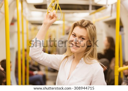 Beautiful blonde young woman holding with right hand inside tube train in London. She wears a white shirt and she is looking at camera smiling. Warm tone applied.
