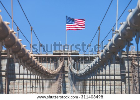 United States Flag at top of Brooklyn Bridge. There is a deep blue sky on background, on foreground there are all the wires of the bridge. Patriotism concept.