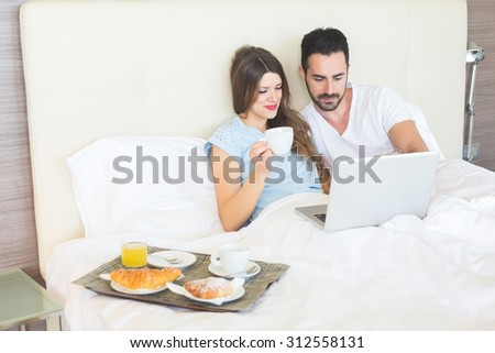 A man and a woman are having breakfast on the bed. They are drinking coffee and juice and eating croissants. They are enjoying relax on holiday but they are also working with computer.