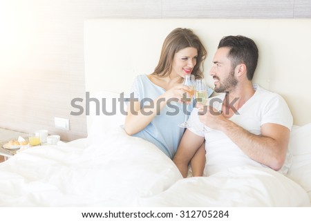 Happy couple making a toast on the bed. It could be on Valentine\'s day or for birthday, they\'re looking each other and smiling. Setting could be luxury home or hotel bedroom.