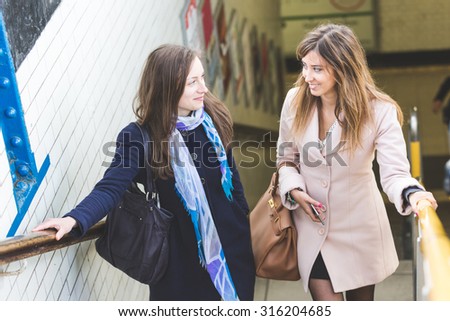 A couple of women are talking while they are coming out from the train station. They are happy, smiling and are going to have a walk together in the city. It\'s winter and both are wearing a coat.