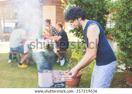 Man cooking meat on the barbecue. He, and the friends of him on background, are all on late twenties. They are eating outdoor on the grass. Everybody is wearing summer clothes.