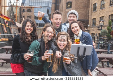 Group of friends taking a selfie with the stick while enjoying a beer at pub in London, toasting and laughing. They are four girls and two boys in their twenties.