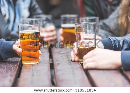 Hands holding glasses with beer on a table at pub in London. A group of friends is enjoying beer time in the city, close up on the glasses.