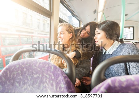 Three women looking out the window from. the bus. They are a mixed group with a caucasian, an asian and a spanish woman. They are friends and they are travelling together by bus.