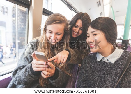 Three women looking a smart phone on the bus. They are a mixed group with a caucasian, an asian and a spanish woman. They are friends and they are travelling together by bus.